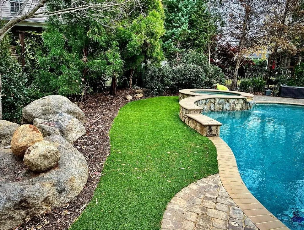 Backyard pool with curved stone edge, built-in spa, and bright green artificial lawn surrounded by trees and rocks.